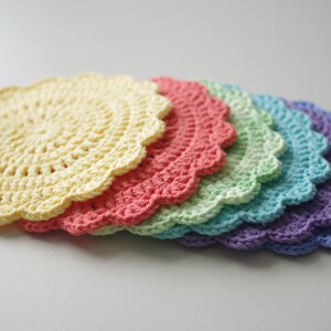 May include: A stack of six crocheted doilies in a rainbow of colors: yellow, coral, light green, teal, and purple. Each doily has a scalloped edge and a circular, open weave pattern. The doilies are arranged in a slight arc.