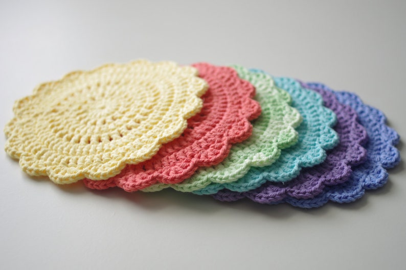 May include: A stack of six crocheted doilies in a rainbow of colors: yellow, coral, light green, teal, and purple. Each doily has a scalloped edge and a circular, open weave pattern. The doilies are arranged in a slight arc.
