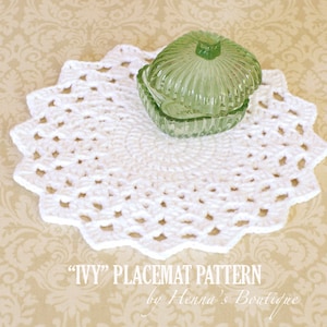 May include: White crochet placemat with a scalloped edge. The placemat is round and has a delicate pattern. A green glass dish is sitting on top of the placemat. "IVY" PLACEMAT PATTERN by Henna's Boutique.