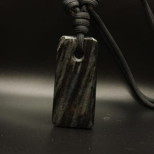 以下が含まれることがあります： 黒とグレーのストライプ模様の長方形ペンダント。黒いコードで吊り下げられています。ペンダントの上部にはコードを通すための穴があります。背景は暗いです。