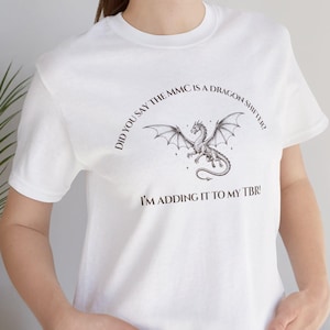 Puede incluir: Camiseta blanca con un diseño de dragón y el texto "DID YOU SAY THE MMC IS A DRAGON SHIFTER? I'M ADDING IT TO MY TBR!". La camiseta está hecha de un material suave.