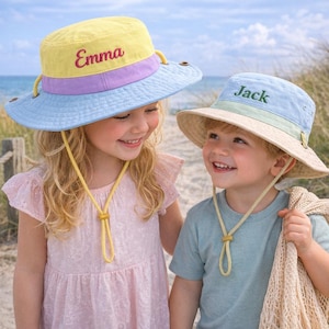 Puede incluir: Dos niños con sombreros de sol personalizados. Los sombreros son en tonos amarillo, azul y beige, con los nombres "Emma" y "Jack" bordados. Los sombreros tienen correas para la barbilla y están diseñados para la protección solar.