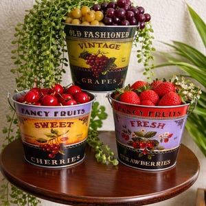 Könnte beinhalten: Drei Metallbehälter im Vintage-Stil, gefüllt mit frischem Obst. Der obere Behälter enthält grüne und lila Trauben, der mittlere Kirschen und der untere Erdbeeren. Jeder Behälter hat ein anderes Farbschema und den Text: "Old Fashioned Vintage Grapes", "Fancy Fruits Sweet Cherries" und "Fancy Fruits Fresh Strawberries".