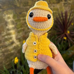 May include: A crocheted duck doll, featuring a yellow hat, waistcoat, and feet, with an orange beak and legs. The duck has black eyes and is wearing a yellow hat and waistcoat with three buttons. The doll is made of white and yellow yarn.