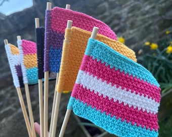 May include: A collection of handmade crochet flags in various colors, including pink, blue, yellow, and purple, each attached to a wooden dowel. The flags represent different LGBTQ+ pride flags, with a focus on the transgender flag.