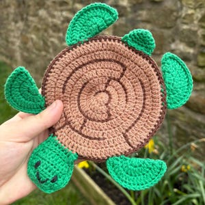 Puede incluir: Un posavasos de tortuga hecho a mano con ganchillo. El posavasos presenta un caparazón marrón con círculos concéntricos y aletas, cabeza y cola verdes. La cara de la tortuga tiene una expresión sonriente. El posavasos se sostiene frente a un fondo borroso.