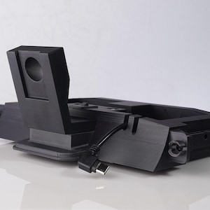 May include: A black charging station with a geometric design, featuring a stand and a curved cable. The station has a rectangular base and a vertical support with a circular aperture. The design is modern and minimalist.