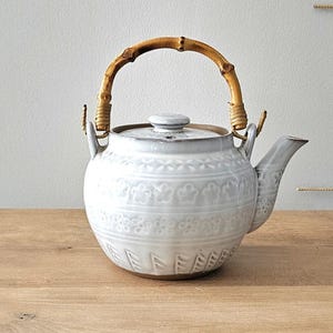 Può includere: Una teiera in ceramica con smalto grigio chiaro e una superficie strutturata con motivi floreali e geometrici in rilievo. La teiera presenta un beccuccio curvo, un coperchio abbinato e un manico curvo realizzato in bambù naturale.