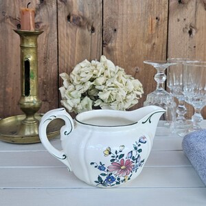 Op de afbeelding: Een crèmekleurig keramieken melkkannetje met een delicaat bloemmotief met roze, gele en blauwe bloemen, geaccentueerd door een dunne groene rand en een gebogen handvat, geplaatst naast een koperen kandelaar en helder glaswerk.