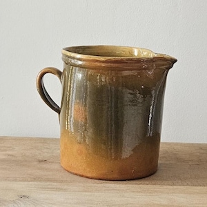 Peut inclure: Une cruche en céramique émaillée, marron et orange, avec une anse, est posée sur une table d'appoint en bois. La table a un tiroir et une étagère inférieure avec des pièces de porcelaine. La cruche est vintage.