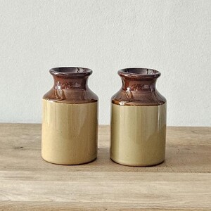 Peut inclure: Deux pots en céramique avec des dessus marron et des corps beiges sont posés sur une table en bois. Les pots ont un design classique et vintage. La table a un tiroir avec une poignée décorative en laiton. Un mur blanc est en arrière-plan.