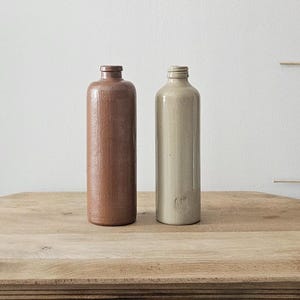 Peut inclure: Deux bouteilles cylindriques sont posées verticalement. La bouteille de gauche est d'une couleur marron mat profond avec une surface texturée. La bouteille de droite est d'une couleur beige clair lisse avec un goulot fileté et une marque.