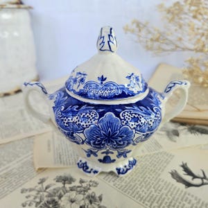 Può includere: Una zuccheriera in ceramica bianca con coperchio, caratterizzata da intricati motivi floreali e a volute blu. Il contenitore presenta due manici laterali, un pomello appuntito sul coperchio e poggia su tre piedini, mostrando un decoro blu e bianco.