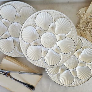 Peut inclure: Trois assiettes à huîtres rondes en céramique blanche, chacune dotée de six empreintes en forme de coquille disposées autour d'un puits central. Les assiettes présentent un motif tressé texturé sur le bord et une finition brillante.