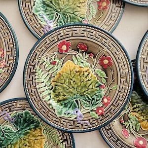Op de afbeelding: Een verzameling ronde keramische borden met een reliëfontwerp van groene bladeren, varens en kleine rode bloemen. Elk bord heeft een bruin midden met een gedetailleerd Grieks sleutelpatroon als rand en een paars lintaccent in reliëf.