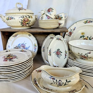 Op de afbeelding: Een crèmekleurige keramische serviesset met een kleurrijk bloemmotief van rode, gele en blauwe bloesems. De collectie bevat een terrine met deksel, een sauskom met onderzetter, diverse borden, kommen en kopjes met bijbehorende schoteltjes.