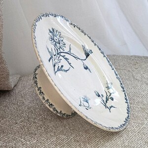 Peut inclure: Un plat sur pied ovale en céramique avec un vernis couleur crème et des illustrations florales bleu foncé. Le bord présente un motif de feuilles bleu délicat, tandis que le centre affiche des dessins botaniques détaillés de fleurs.