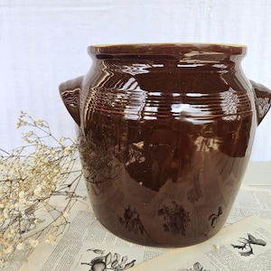 Może przedstawiać: Duży, ciemnobrązowy, glazurowany garnek ceramiczny z szerokim otworem, grubym brzegiem i dwoma bocznymi uchwytami. Zewnętrzna powierzchnia jest gładka i odblaskowa, z poziomymi żebrowanymi detalami wokół górnej części szyjki naczynia.