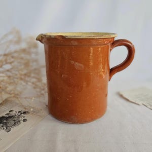 Puede incluir: Una jarra cilíndrica de cerámica con un esmalte brillante de color marrón anaranjado intenso. El recipiente presenta un asa curva sencilla en el lado derecho, un pequeño pico vertedor y un borde de color crema claro. La superficie muestra texturas.