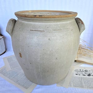 Pode incluir: Um pote de grés grande e redondo em cinza claro, com duas alças laterais e uma borda larga não esmaltada em tom bronzeado. A superfície externa apresenta uma textura desgastada com sutis sulcos horizontais e pequenas manchas escuras.