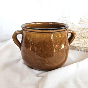 Op de afbeelding: Een ronde, bruine keramische pot met een glanzende afwerking en twee handvatten aan de zijkanten. De pot heeft een brede opening met een donkerbruine rand en een gladde, gespikkelde textuur over het hele lichaam, met een functioneel ontwerp.