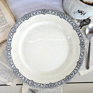 Könnte beinhalten: Ein runder, cremeweißer Keramikteller mit einem dekorativen Rand, der ein sich wiederholendes dunkelblaues Blumenmuster in ovalen Rahmen aufweist. Der Teller hat eine glatte, glänzende Oberfläche und einen breiten Rand mit klassischem Design.