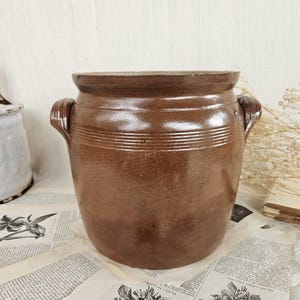 Op de afbeelding: Een bruin geglazuurde keramische pot met twee zijhandvatten en horizontale ribbeldetails rond het bovenste gedeelte. Het vat heeft een brede opening en een afgeronde basis, met een gladde, glanzende afwerking aan de buitenkant.