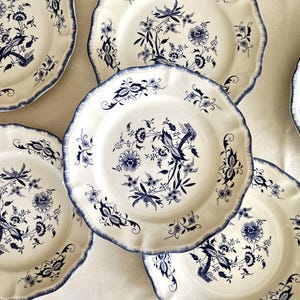 Może przedstawiać: Zestaw pięciu kremowych talerzy ceramicznych z falistym brzegiem i niebieskim wzorem kwiatowym. Każdy talerz posiada centralny motyw botaniczny ze szczegółowymi niebieskimi łodygami, liśćmi i kwiatami, uzupełniony niebieskim wykończeniem brzegu.