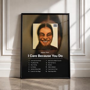 May include: A framed poster featuring the Aphex Twin album I Care Because You Do. The artwork displays a portrait of a smiling person with long dark hair, a barcode, and a tracklist numbered 1 through 12. The background is a dark gradient.