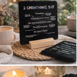 Puede incluir: Una pizarra negra con el texto "2 CORINTIOS 5:17" y un mensaje escrito a mano. La pizarra está apoyada en un pequeño soporte de madera. Una pila de tarjetas negras con texto blanco está en primer plano.