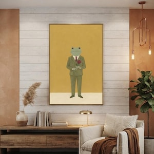 May include: A framed wall art print featuring a green frog dressed in a tan suit with a red tie, holding a red rose. The frog stands against a solid mustard yellow background with a white horizontal band at the bottom of the rectangular frame.