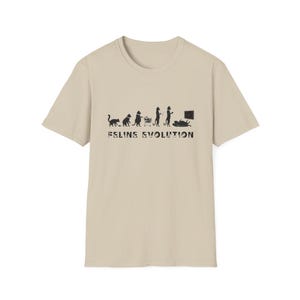 May include: A beige t-shirt with a black graphic depicting cat evolution. The design shows cat silhouettes, evolving from a cat to a cat with a shopping cart, and finally, a cat watching TV. The words "Feline Evolution" are printed below.