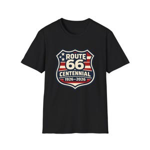 Puede incluir: Camiseta negra con un diseño del Centenario de la Ruta 66. El gráfico incluye una forma de escudo con las palabras "Route 66 Centennial 1926-2026" en rojo, blanco y azul. La camiseta está hecha de un material suave.