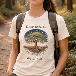 Pode incluir: Uma camiseta creme com uma ilustração detalhada de uma árvore com raízes profundas e uma copa cheia sob um céu estrelado. O texto diz "DEEP ROOTS WILD SOUL" com a frase "grounded enough to grow - free enough to bloom."