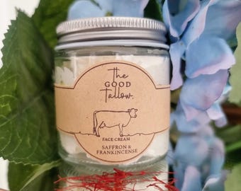 Op de afbeelding: Een klein glazen potje met een zilveren deksel, gevuld met witte crème. Een bruin etiket leest "The Good Tallow" met een koe-illustratie en "Face Cream Saffron & Frankincense". De pot is omgeven door blauwe bloemen en groene bladeren.