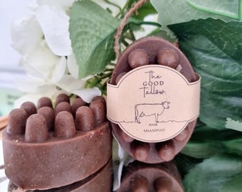 Op de afbeelding: Twee bruine, ovale haarshampoor bars met een getextureerd oppervlak. Eén bar heeft een label met "The Good Tallow", een koe-illustratie en "Hair Shampoo". De bars worden gepresenteerd op een reflecterend oppervlak met bloemaccenten.