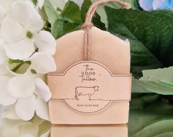 Op de afbeelding: Een lichtbeige bodyzeep met een bruin papieren label met de tekst "The Good Tallow" en een koe-illustratie. De zeep is vastgebonden met een natuurlijke touwtje. De woorden "Body Soap Bar" staan op het label.