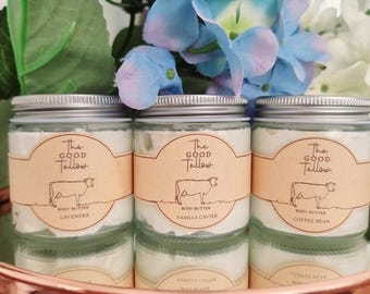 Op de afbeelding: Drie glazen potten body butter met zilveren deksels en etiketten met de tekst "The Good Tallow". De etiketten tonen een koe-illustratie en de smaken: Lavendel, Vanille Caviar en Koffieboon. De potten zijn gerangschikt op een koperkleurig dienblad.