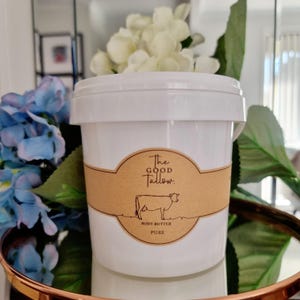 May include: A white plastic tub of "The Good Tallow" body butter. The label is brown with a cow illustration. The tub is surrounded by blue and white flowers and greenery, sitting on a copper-coloured tray.