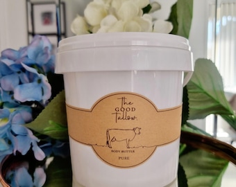 Op de afbeelding: Een witte plastic emmer met "The Good Tallow" body butter. Het etiket is bruin met een koe-illustratie. De emmer is omgeven door blauwe en witte bloemen en groen, en staat op een koperkleurig dienblad.