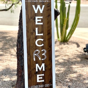 May include: A rectangular wooden welcome sign with a dark brown centre and white border. The word "WELCOME" is displayed in large white letters, with "R3" in the middle. The bottom reads "ESTABLISHED 2021". Coordinates are at the top.