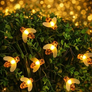 May include: A string of bee-shaped fairy lights with a warm yellow glow, nestled among green foliage. The lights have translucent wings and a detailed body, creating a whimsical and inviting ambiance. The background is blurred with additional bokeh lights.