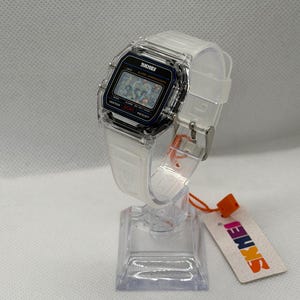 May include: A digital wristwatch with a clear, transparent resin case and a matching white translucent silicone strap. The rectangular watch face features a black border, a digital display, and the text SKMEI, ALARM CHRONOGRAPH, and WATER 50M RESIST.