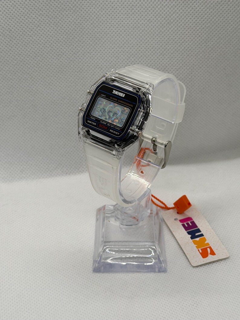 May include: A digital wristwatch with a clear, transparent resin case and a matching white translucent silicone strap. The rectangular watch face features a black border, a digital display, and the text SKMEI, ALARM CHRONOGRAPH, and WATER 50M RESIST.