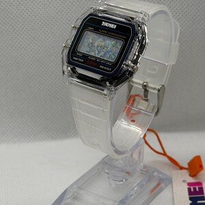 May include: A digital wristwatch with a clear transparent plastic case and a white silicone strap. The watch face features a black rectangular display with the brand name SKMEI, the text ALARM CHRONOGRAPH, and WATER 50M RESIST printed in white.