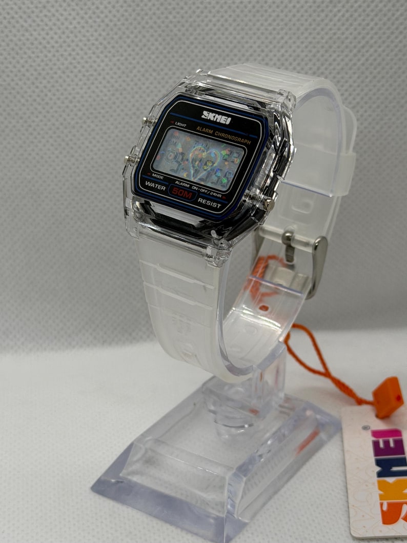 May include: A digital wristwatch with a clear transparent plastic case and a white silicone strap. The watch face features a black rectangular display with the brand name SKMEI, the text ALARM CHRONOGRAPH, and WATER 50M RESIST printed in white.