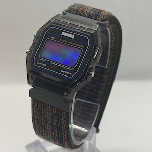 May include: A digital wristwatch with a translucent dark gray plastic case and a rectangular face. The display features the text SKNEI, ALARM CHRONOGRAPH, and WATER 50M RESIST. It has a woven fabric strap in a dark blue and orange textured pattern.