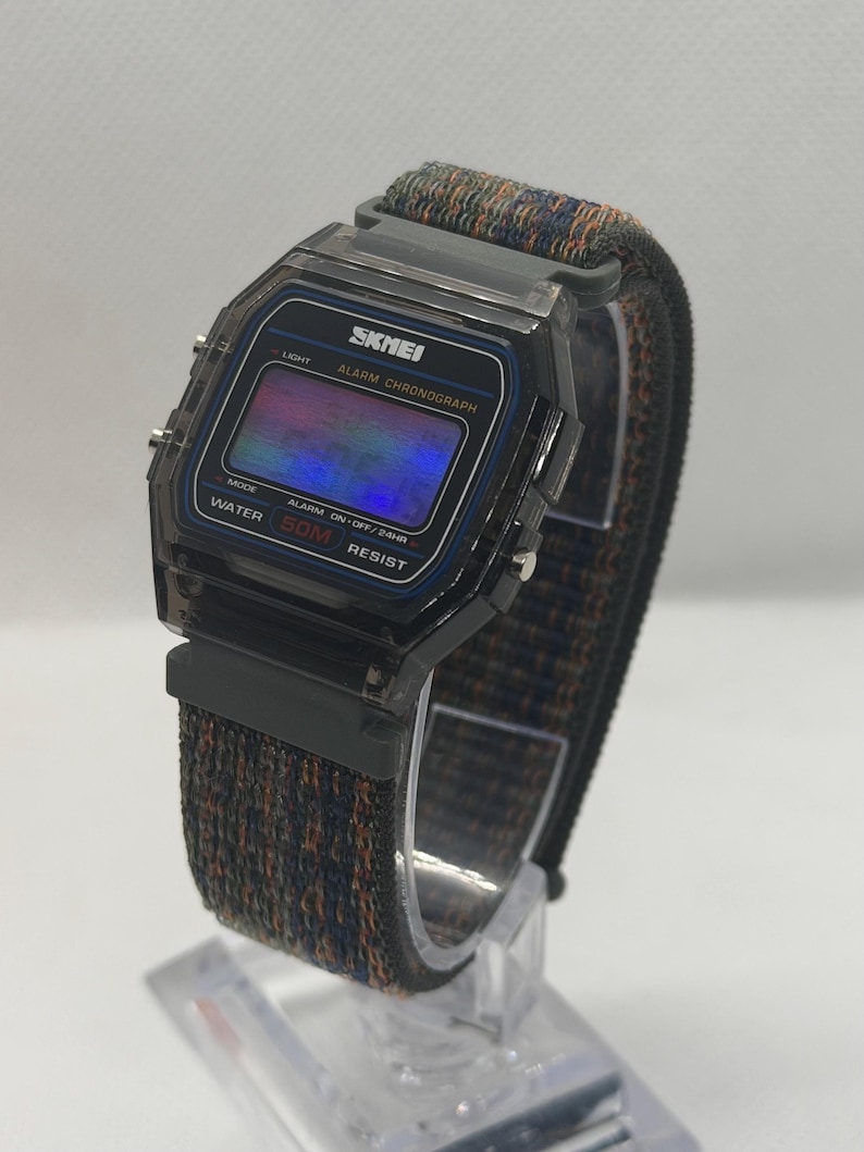 May include: A digital wristwatch with a translucent dark gray plastic case and a rectangular face. The display features the text SKNEI, ALARM CHRONOGRAPH, and WATER 50M RESIST. It has a woven fabric strap in a dark blue and orange textured pattern.