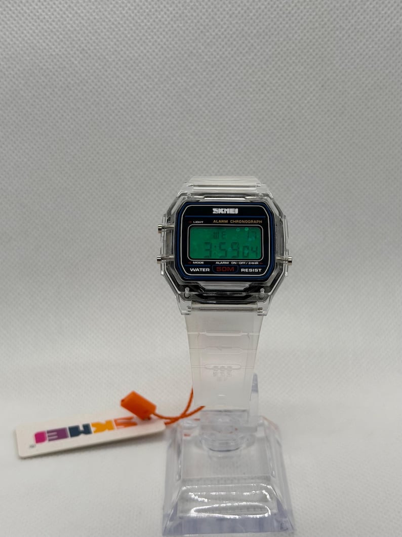 K&ouml;nnte beinhalten: Transparente Digitaluhr mit rechteckigem Zifferblatt und transparentem Armband. Das Zifferblatt zeigt die Uhrzeit in gr&uuml;nen Ziffern an. Die Uhr hat den Markennamen "SKMEI" auf dem Zifferblatt. Ein kleines Etikett mit dem Markennamen ist angebracht.