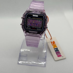 Puede incluir: Un reloj digital lavanda transparente con una esfera negra que muestra la hora. El reloj tiene una correa lavanda a juego y est&aacute; sobre un soporte de pl&aacute;stico transparente. La marca "SKMEI" es visible en la esfera y la etiqueta.